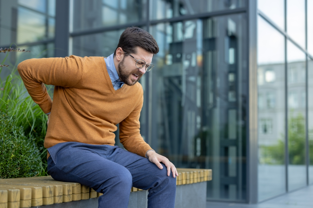 Young tired male businessman sitting on a bench outside an office center and holding his hand behind his back